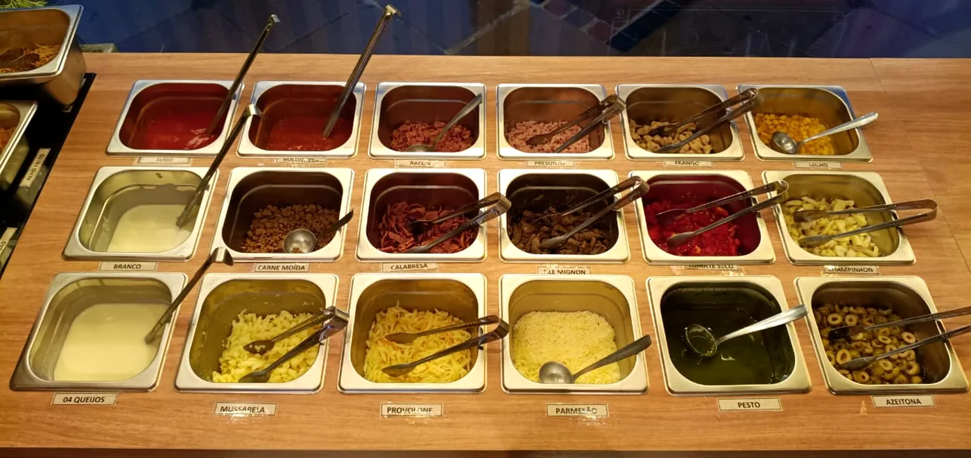 Bancada de acompanhamentos da Massita em Gramado, com cubas de molhos, carnes, queijos e legumes identificados por etiqueta